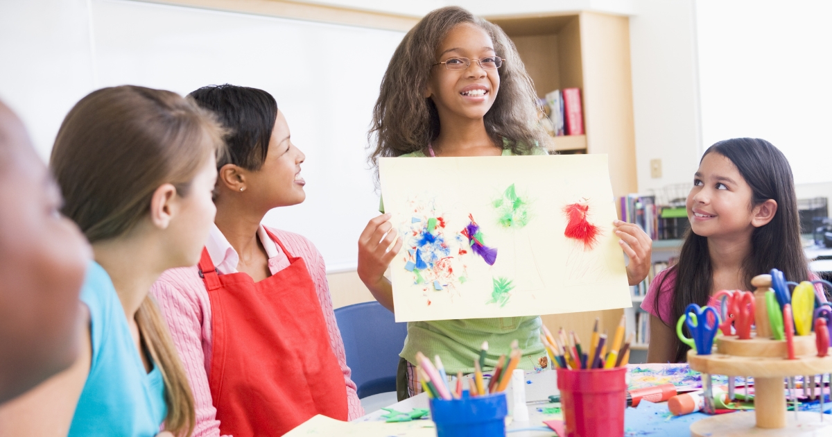 Smiling child presenting her artwork in a creative classroom, surrounded by peers and art supplies, promoting creativity and self-expression in children.